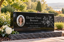 Black granite memorial with a color ceramic portrait, fictional name and dates, engraved remembrance inscription, and floral design in a garden.